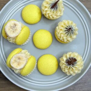 Displayed on a round gray plate, six Banana Nutella Macarons topped with banana slices accompany cream-swirled cupcakes with chocolate accents. Perfect for any UK gathering, this delightful selection rests on a wooden surface, inviting you to indulge in these exquisite treats.