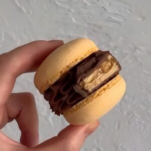 A hand holds a Snickers Macaron with yellow shell and chocolate-nut cream filling, perfect for those seeking indulgence. The macaron's light, textured surface reflects Scotland's quaint charm.