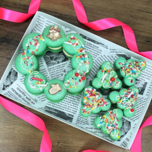A delivery box contains Kinder Bueno Macarons in holiday-themed designs, shaped like wreaths and trees. They rest on newspaper, ready to delight recipients across the UK. A pink ribbon around the box on a wooden surface adds a final touch of charm.