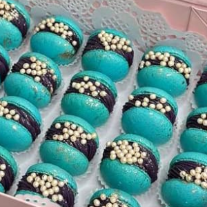 A box of Blueberry Macarons features blue shells with black filling, adorned with white pearls and arranged on lace-patterned paper. These elegant treats add a touch of fine Scottish craftsmanship to any UK celebration.