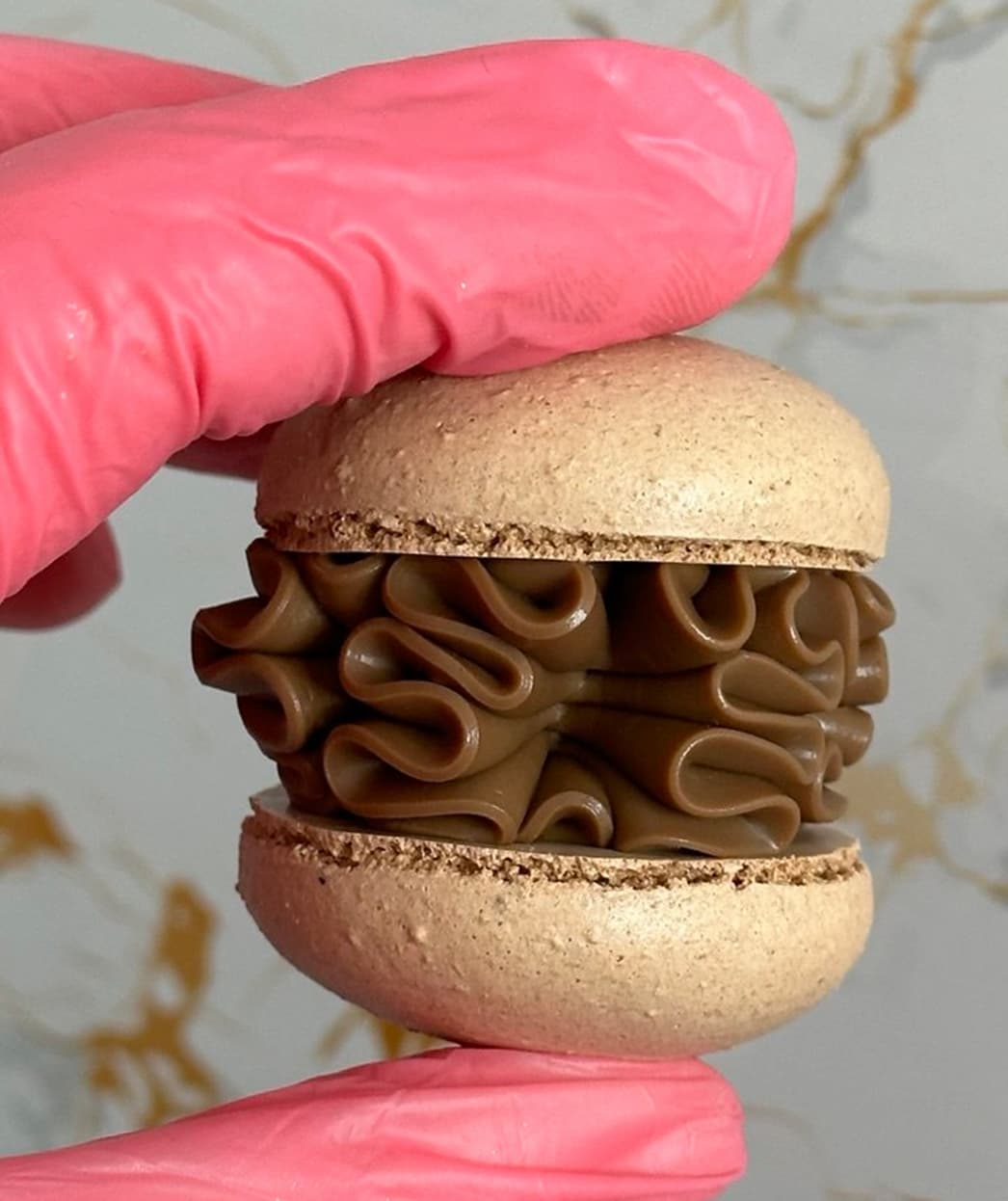 A pink-gloved hand holds a Latte Macaron, packed with ruffled chocolate filling. The elegant light gray and gold marble background adds a touch that would elevate any delivery service, reminiscent of exquisite Scottish creations.