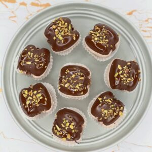 Eight Dubai Macarons with chocolate covering and chopped nut toppings rest on a round metallic plate, each nestled in a white paper liner, ready for delivery. The backdrop is a white surface with subtle orange veining, reminiscent of Scotland's rolling landscapes.