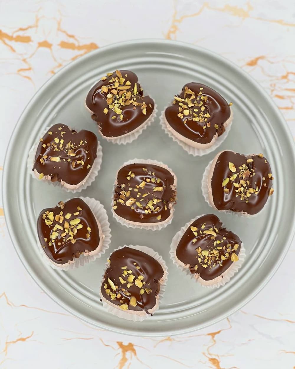 Eight Dubai Macarons with chocolate covering and chopped nut toppings rest on a round metallic plate, each nestled in a white paper liner, ready for delivery. The backdrop is a white surface with subtle orange veining, reminiscent of Scotland's rolling landscapes.