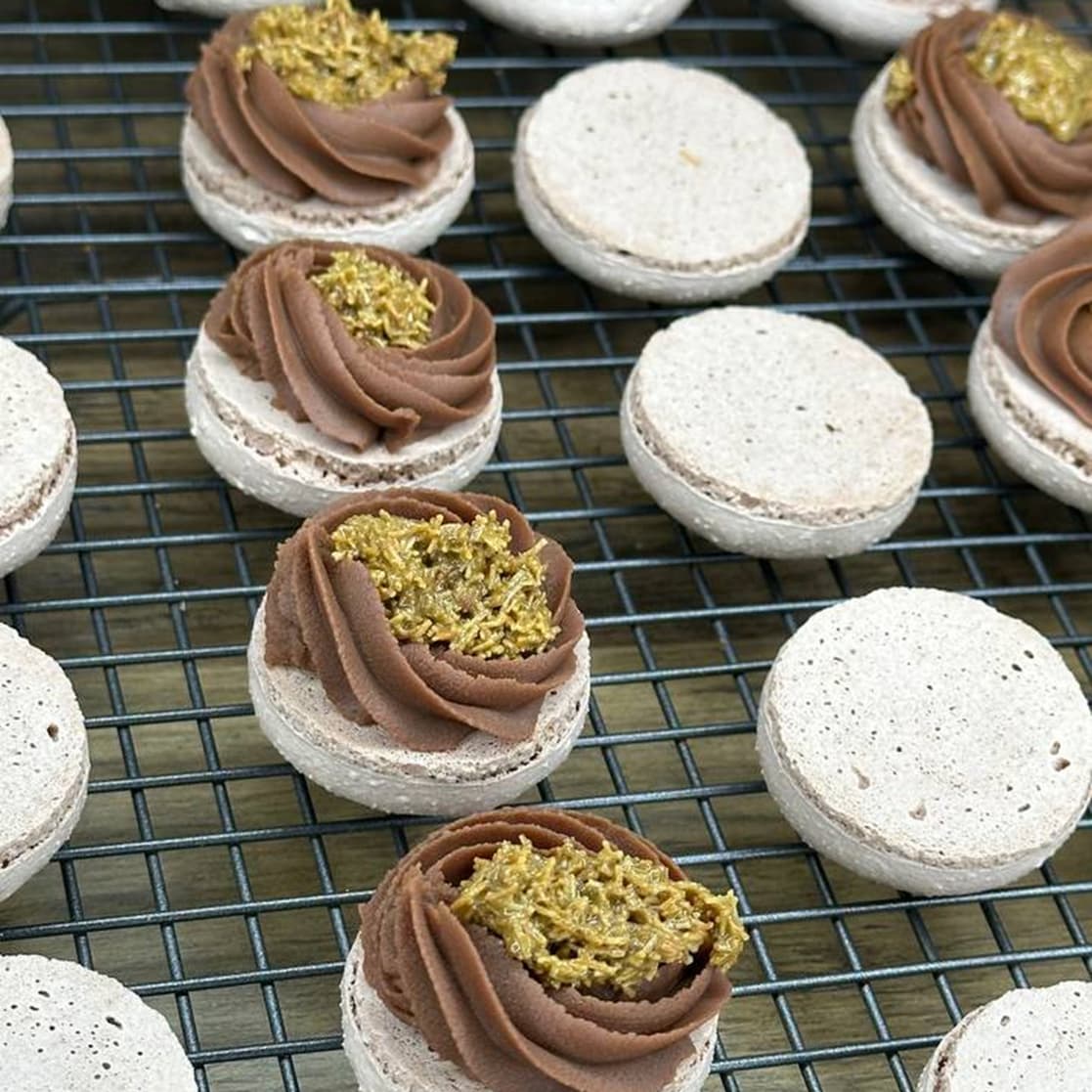 Unfilled Dubai Macarons rest on a cooling rack, some adorned with swirls of chocolate cream and gold sprinkles, ready for delivery across Scotland.