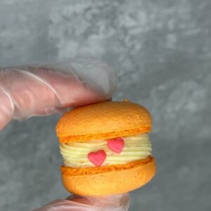 A hand in a transparent glove holds an Orange Passion Fruit Macaron with creamy filling, adorned with two small pink heart decorations against a textured gray background, reminiscent of delicate UK confections.