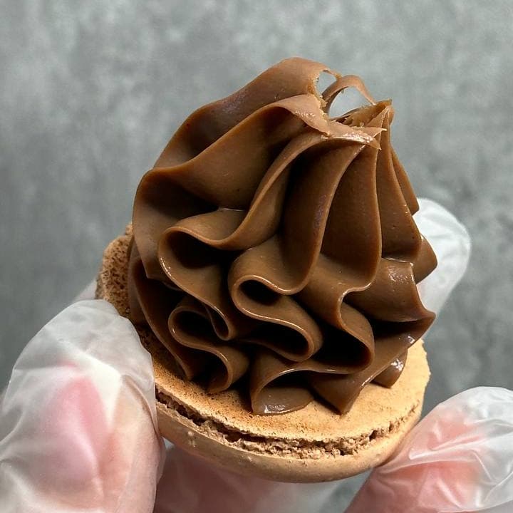 A clear-gloved hand holds a Latte Macaron topped with rich chocolate frosting. The macaron's light brown shell has slightly cracked edges, set against a blurred gray background.