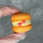 A gloved hand holds an orange macaron filled with cream, adorned with two pink heart-shaped decorations. Perfect for a custom gift, this delightful treat stands out against a blurred gray background, showcasing the charm of custom macarons.