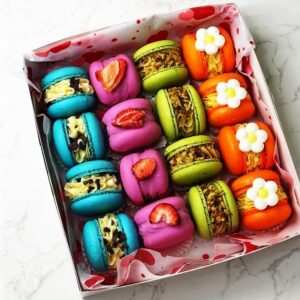 A Pier-Re-Her-Me Macarons Box sits on marble, displaying vibrantly arranged macarons: blue with chocolate chips, pink with strawberry slices, green with nuts, and orange topped with white flower designs. The interior of the box is lined with a heart pattern.