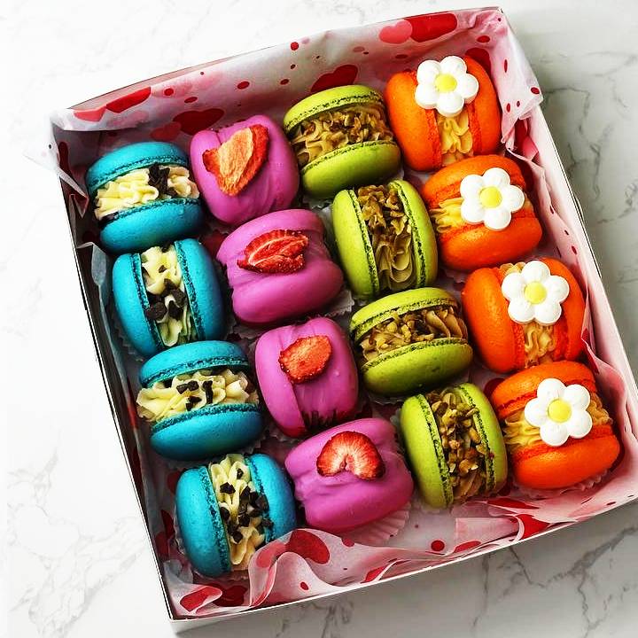 A Pier-Re-Her-Me Macarons Box sits on marble, displaying vibrantly arranged macarons: blue with chocolate chips, pink with strawberry slices, green with nuts, and orange topped with white flower designs. The interior of the box is lined with a heart pattern.