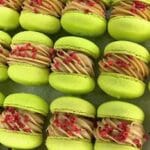 A close-up of rows of custom lime green macarons filled with light brown cream. The filling is topped with small red pieces, possibly fruit or candy, all perfectly arranged as if awaiting placement in a decadent macarons box.