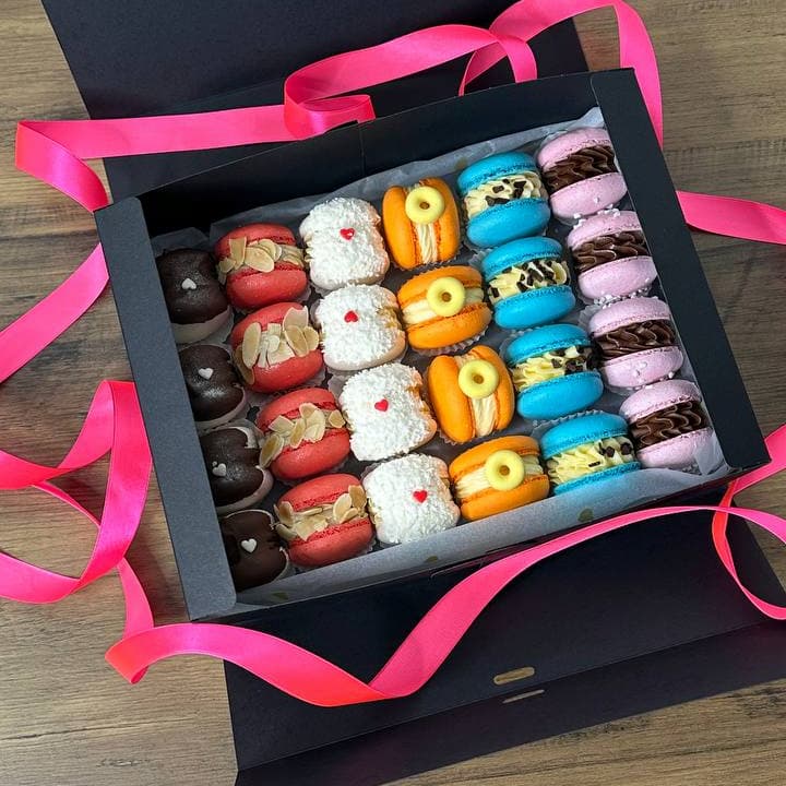 A custom box of assorted macarons sits open on a wooden surface, adorned with pink ribbons. The vibrant macarons are arranged in neat rows, showcasing an array of colors like red, white, orange, blue, and purple.