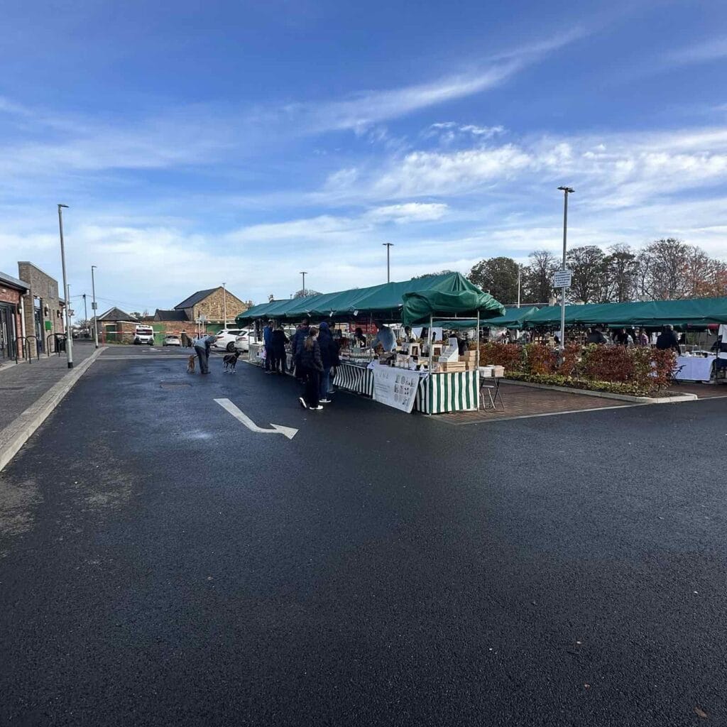 Roswell Market outdoor market with green canopies, vendor stalls, and people shopping on a wide paved area under a partly cloudy sky.