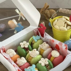 Eine Osterblüten-Macaron-Schachtel steht neben der Osterdekoration und einem gelben Eimer, der mit weißer Schokolade gefüllt ist.