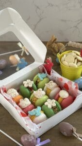 A box of colorful macarons decorated with flowers and butterflies sits on a wooden table with Easter eggs, a yellow pail, and rabbit figurines.