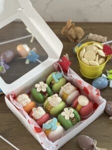 A white box with heart-patterned paper holds assorted colorful macarons. Nearby are Easter eggs, a yellow pail of white chocolate, and rabbit figurines.