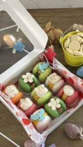 A box of assorted decorated macarons in pastel colors sits on a wooden table with Easter-themed decorations around it.
