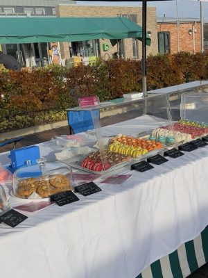 A Roswell Market stall with trays of colorful macarons, cookies, brownies, and baked goods on a striped tablecloth outdoors.