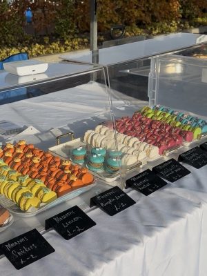 A display of colorful macarons at Roswell Market, arranged in rows under clear cases, each flavor labeled with a small black sign.