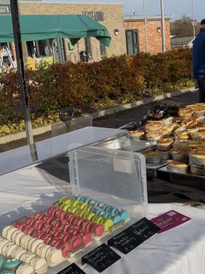 Macarons and pies displayed on a Roswell Market stall table with price tags, framed by outdoor buildings and autumn foliage in the background.