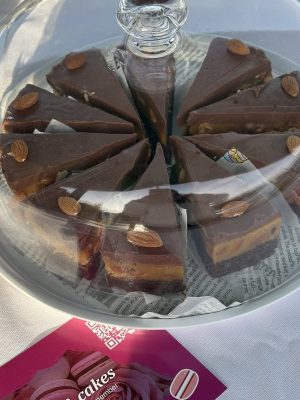 Slices of chocolate caramel cake with almond toppings at Roswell Market, displayed under a glass dome beside a pink business card.
